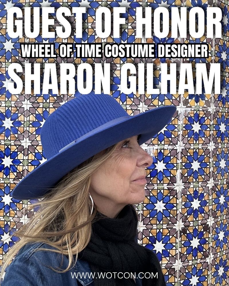 an image with the caption guest of honor wheel of time costume designer Sharon Gilham, WoTCon.com. The image features a blonde woman, with a wide-brimmed blue hat, facing to the side and looking up with a grin on her face. She’s wearing a black scarf and jean jacket, and behind her is mosaic tile in a blue flower pattern with a gold outline