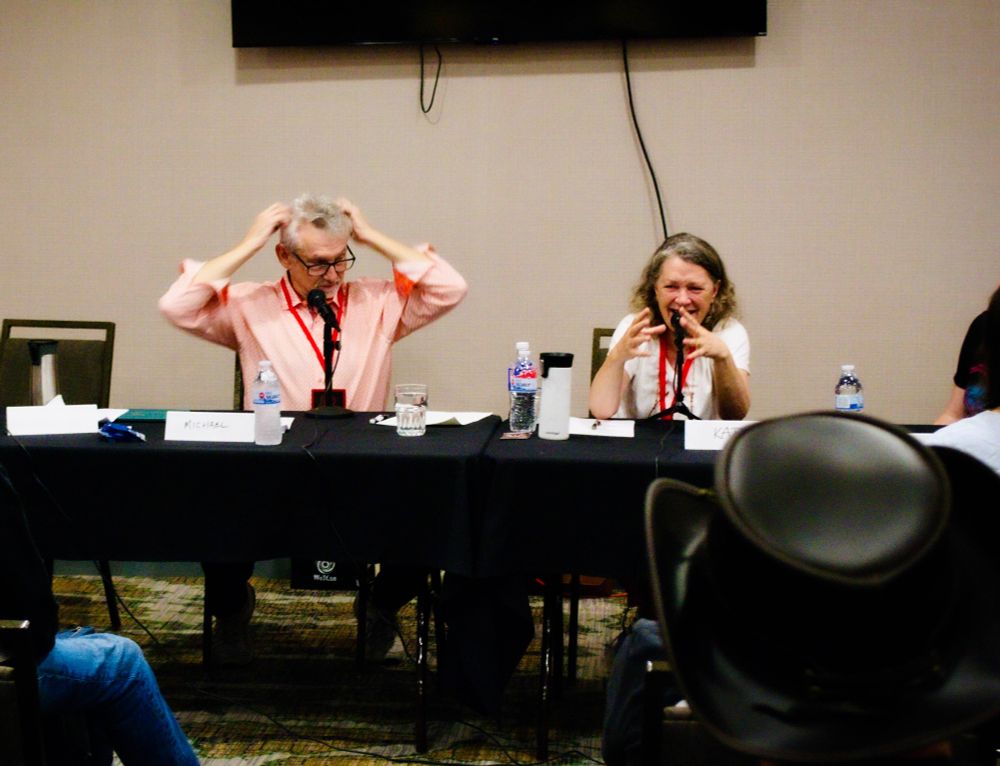 Michael Kramer—a smiling man with a grey/white beard and hair and arms raised, with his hands grasping his hair; and Kate Reading—a woman with shoulder-length wavy sandy-brown hair talking into a microphone with her hands stretched out as if to encircle the microphone. They are sitting at a table as WoTCon panelists.