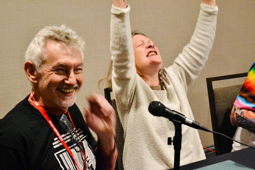 Michael Kramer—a sitting and smiling man with a grey/white beard and hair with on hand extended toward his face; and Kate Reading—a seated woman with shoulder-length wavy sandy-brown hair, reaching toward the ceiling with both arms with an expression as if she’s yelling “Yes!” There are mics in front of them both as they’re doing a panel at WoTCon.