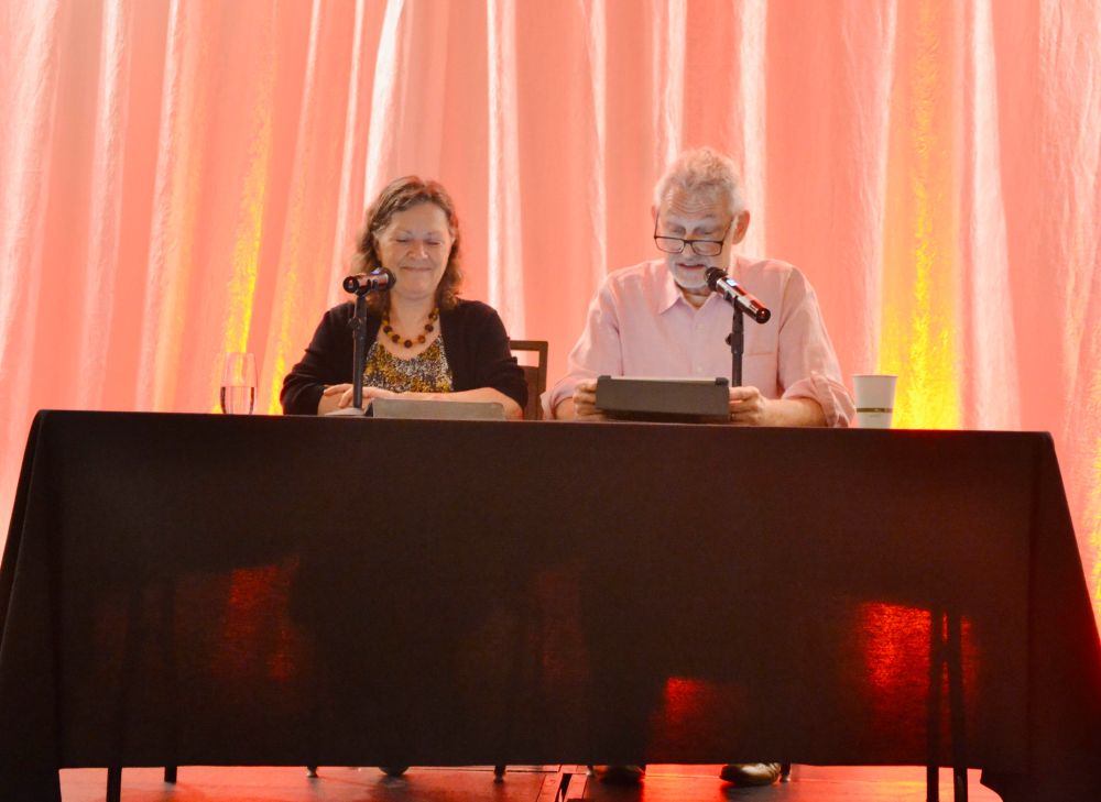 Michael Kramer—a man with a grey/white beard and hair and reading glasses; and Kate Reading—a grinning woman with shoulder-length wavy sandy-brown hair. They are on the main stage at WoTCon, sitting at a table holding iPads for a reading.