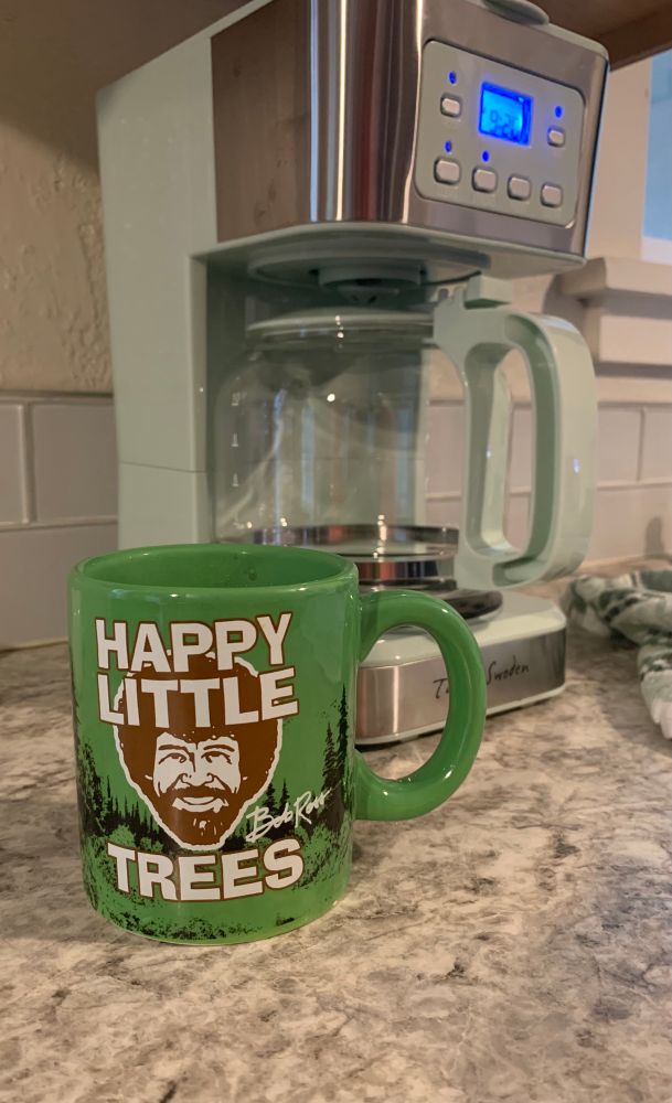 Bob Ross smiling on a green coffee mug with “Happy Little Trees” quote. 