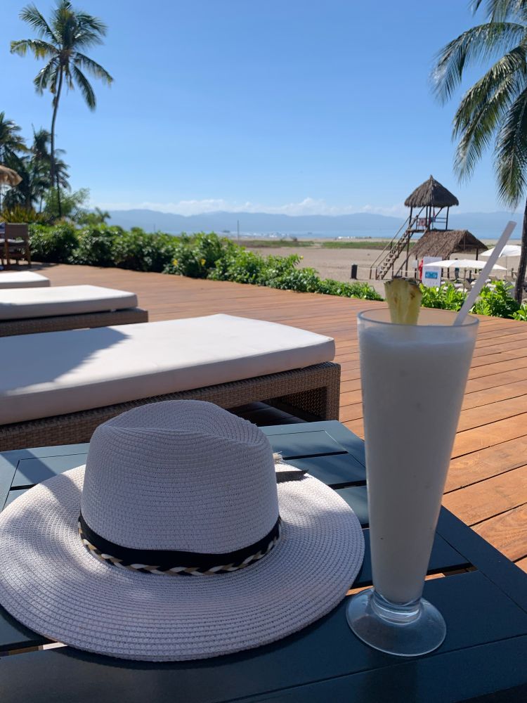 Sun hat on a table next to a frozen drink with the beach and umbrellas in the background. 