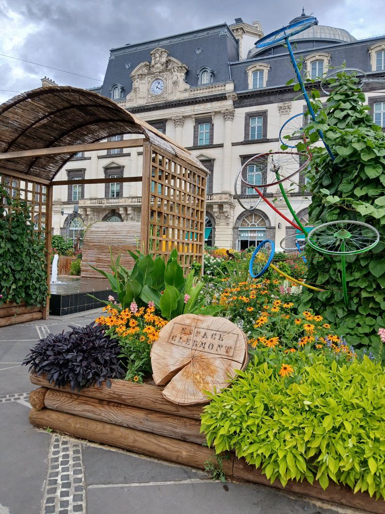 Entrée d'un jardin urbain appelé "Espace Clermont", d'après ce qui est marqué sur la souche qui en marque l'entrée. 
On y voit des massifs de fleurs et une fontaine dans le fond après une arche en bois.