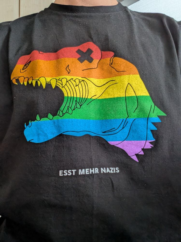 Schwarzes Band-Shirt der Band Treptow mit einem regenbogenfarbenen T-Rex-Kopf. Darunter der Satz "Esst mehr Nazis"