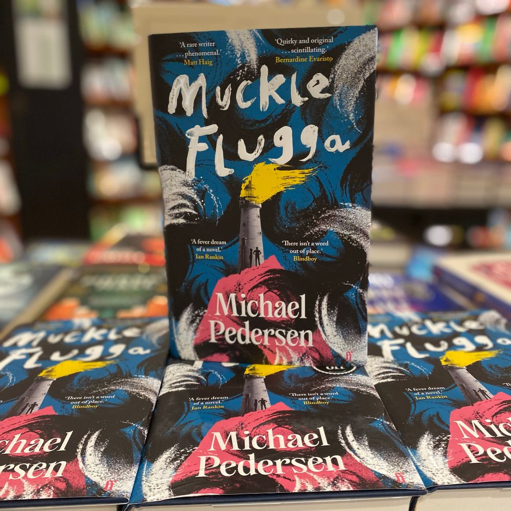 A blurred bookshop background. In centre frame is a table on which a hardback copy of Muckle Flugga by Michael Pedersen stands atop three other copies. The painterly-style cover has white text with and the image shows a lighthouse atop a pink rock, surrounded by sweeps of colour (white, blue and black) that represent waves and the darkness. The lighthouse emits a streak of yellow light and silhouettes of people stand at its base.