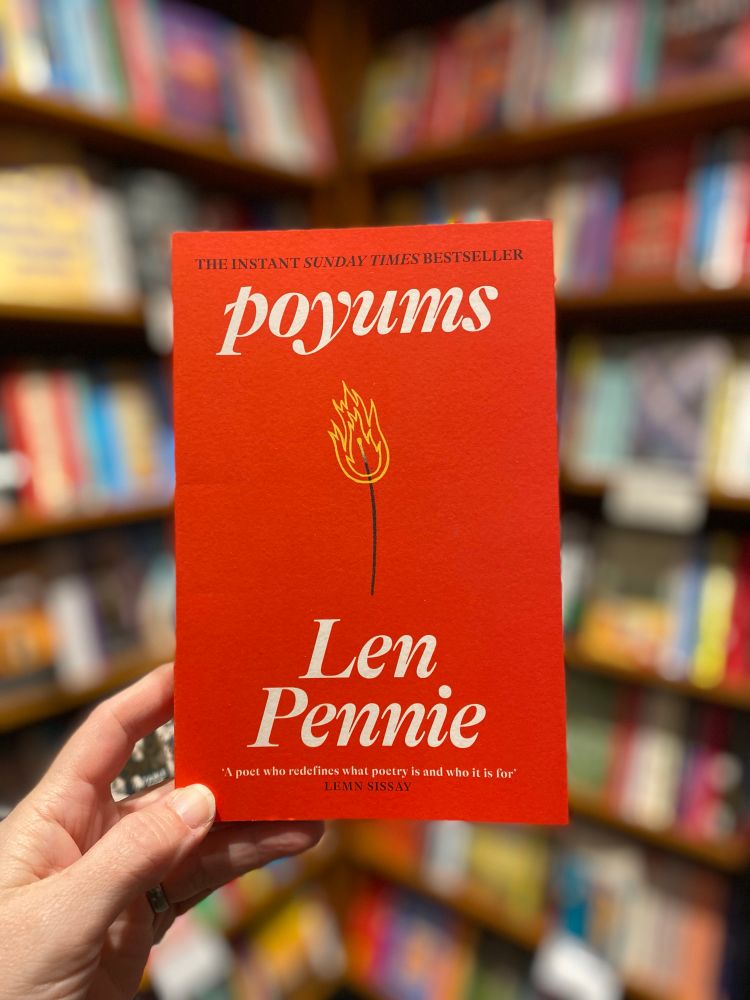 In front of a blurred bookshelf background a hand holds a red paperback book - Poyums by Len Pennie. There is a minimalist illustration on the cover of a burning match, in black and yellow. The title and author text is white and a quote from Lemn Sissay reads ‘A poet who redefined what poetry is and who it is for’.