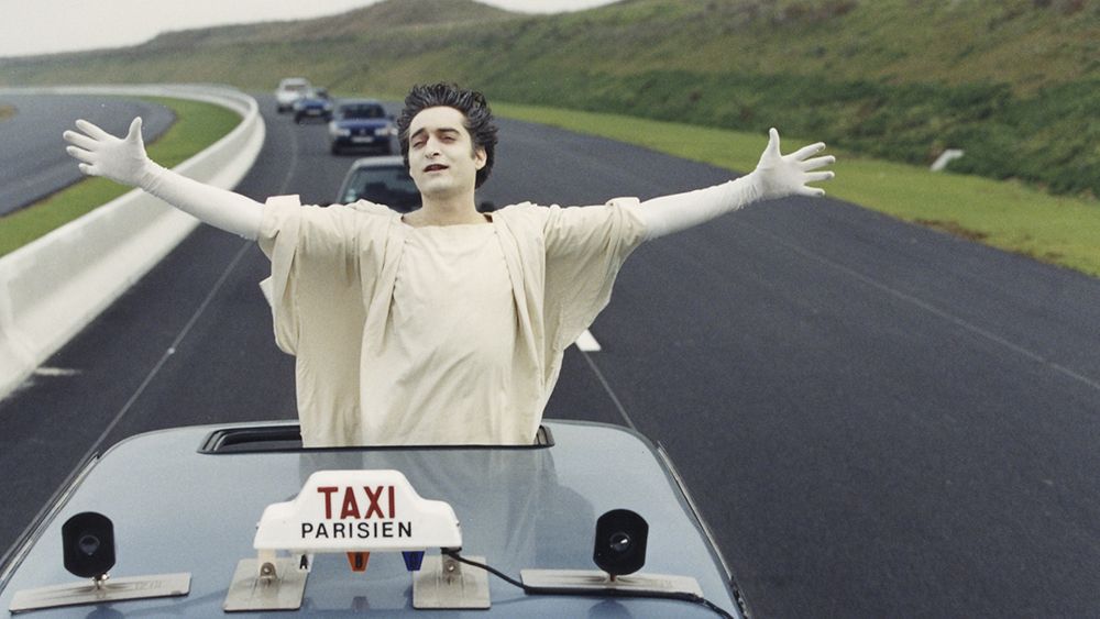 Jean-Claude (Mathieu Demy) les bras écartés, le corps dépassant du toit ouvrant d’une voiture sur l’autoroute.