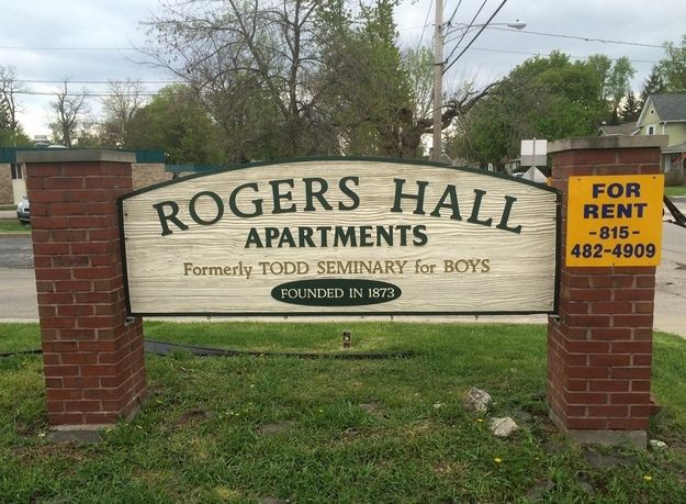 A sign for the Rogers Hall Apartments in Woodstock, IL. The sign reads, "Rogers Hall Apartments. Formerly Todd Seminary for Boys. Founded in 1873." A "For Rent" sign with a local phone number hangs to the right on a red brick post. 