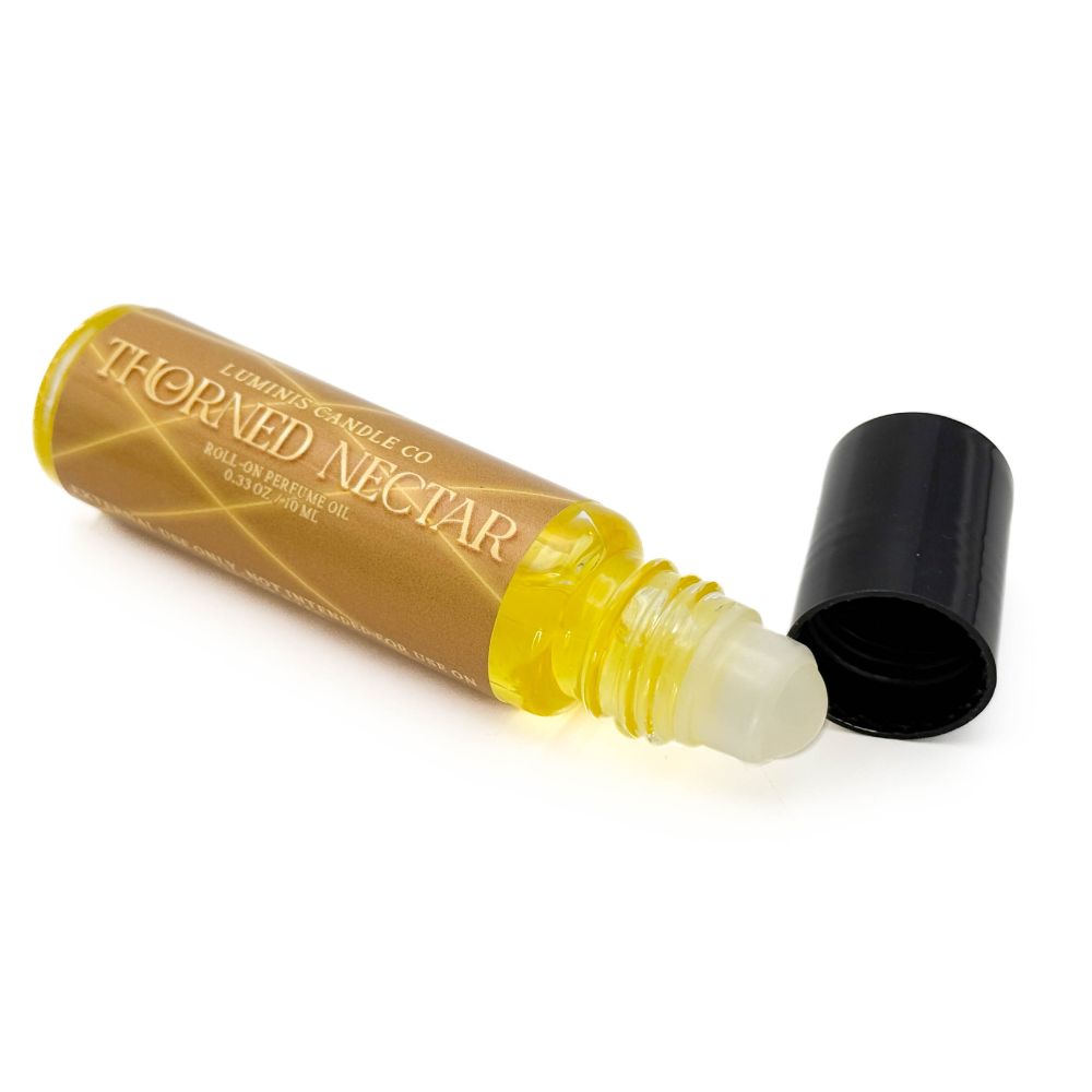 A roll-on vial containing a saturated yellow oil. The vial's black cap is removed and set to the side. The vial has a dark gold label with golden threads criss-crossing it, and gold text reads "Thorned Nectar".
