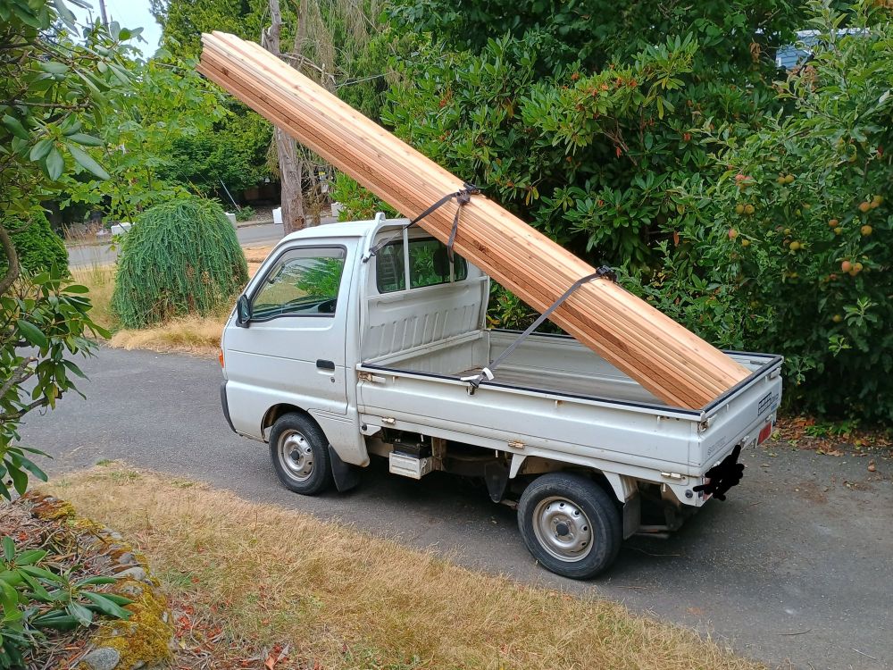 kei truck with a large load of very long lumber in the bed hanging over the cab by a lot