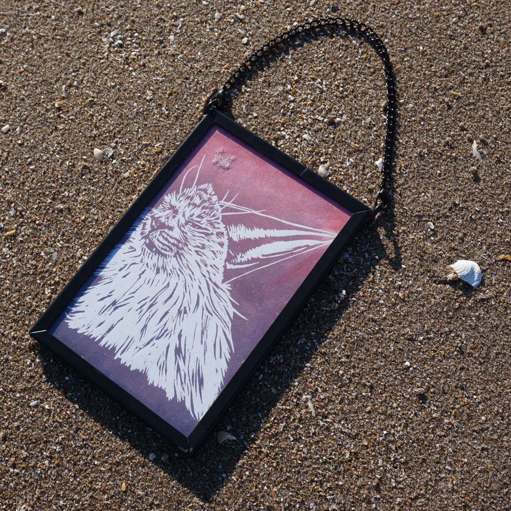 A frame on sand with art of a cat with a party hat on