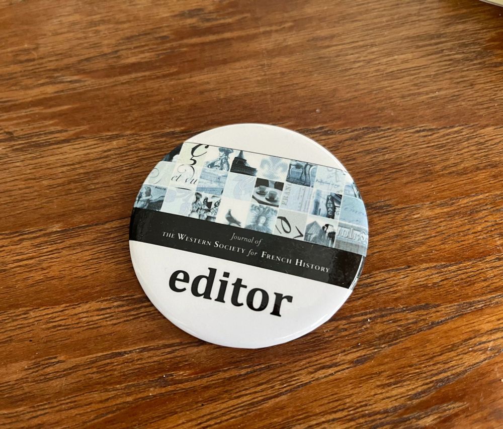 A round badge on a wooden table- editor is written in lower caps at the bottom in black against a white background and the logo for JWSFH above.