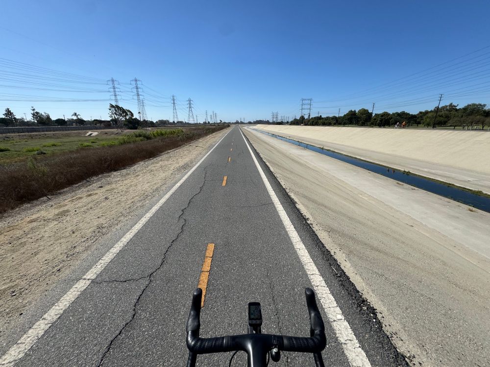 A pair of road bike handlebars pointed down the Los Angeles river bike path. To the right is a steep drop off to the concrete channel of the river, to the left a gravel drop off.