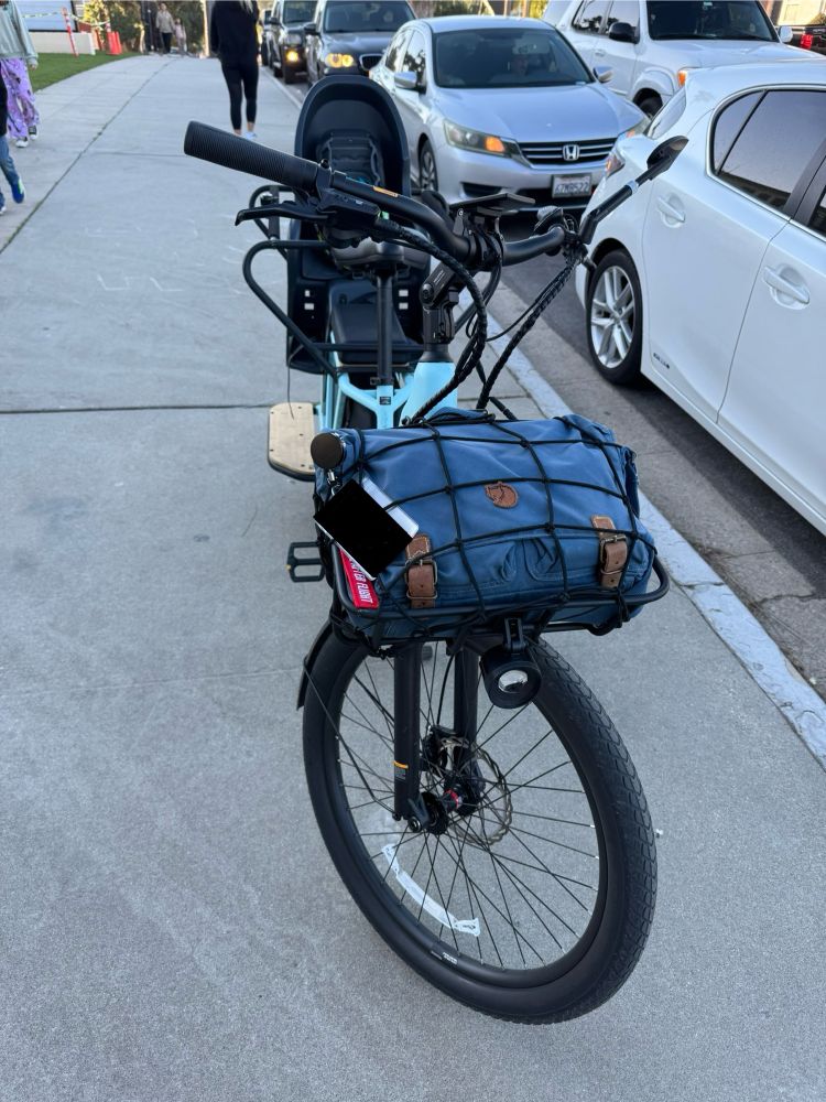 A cargo bike, the weird trick alluded to in the skeet, sits parked on the sidewalk, having recently dropped off a kid at school. There’s a long line of cars in the background, posing no impediment to our brave two wheeled steed.