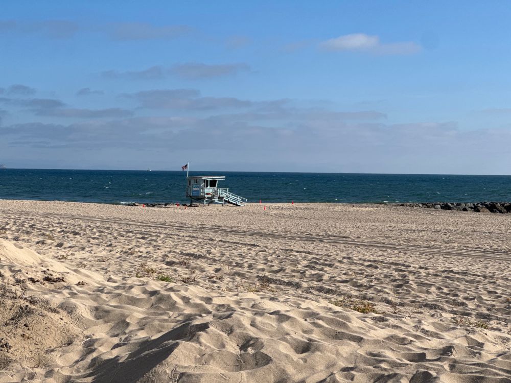 A lone lifeguard tower stands amongst the sand, watching over any who venture into the ocean. It’s light blue and squat, enclosed with windows all around and a ramp leading down to the sand. A small American flag flies from a flagpole on it.
Behind it, the Pacific Ocean stretches away to the horizon.
Above it, blue skies with the beginnings of the marine layer clouds beginning to form on the ocean, ready to roll in with the evening breeze.
Before it, a vast expanse of yellow sand.