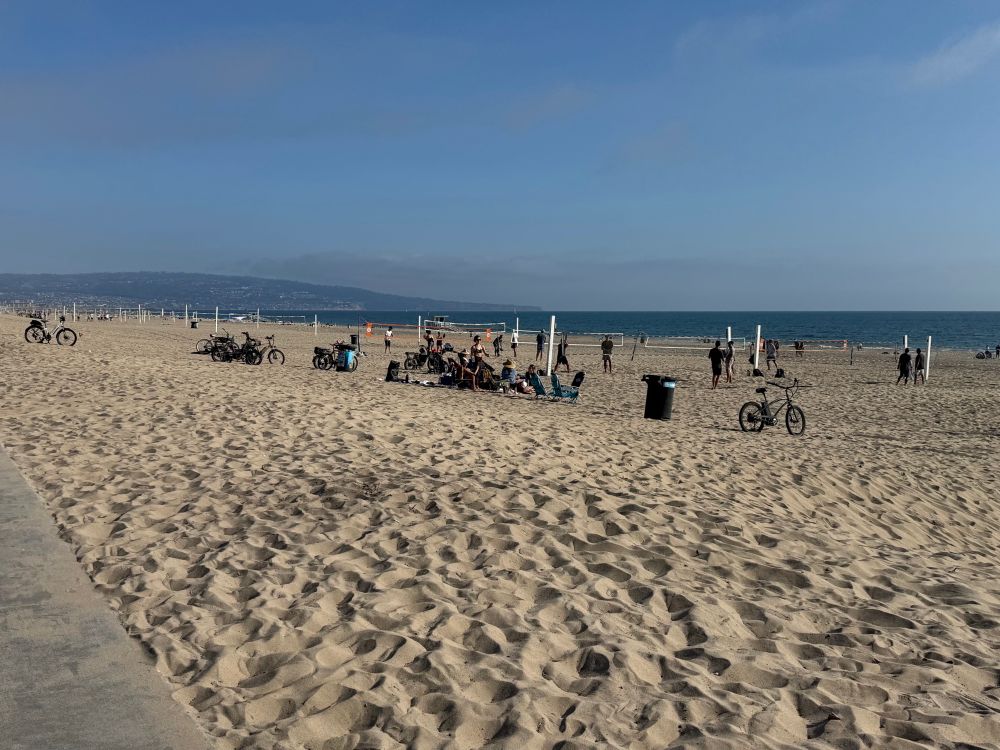 A picture of Manhattan Beach, in Los Angeles. The sun is shining, the day is perfect, there’s a very blue sky, yellow sand, volleyball nets, and a good selection of people playing volleyball and hanging out. There’s a ton of people in beach chairs and bikes parked on the sand.