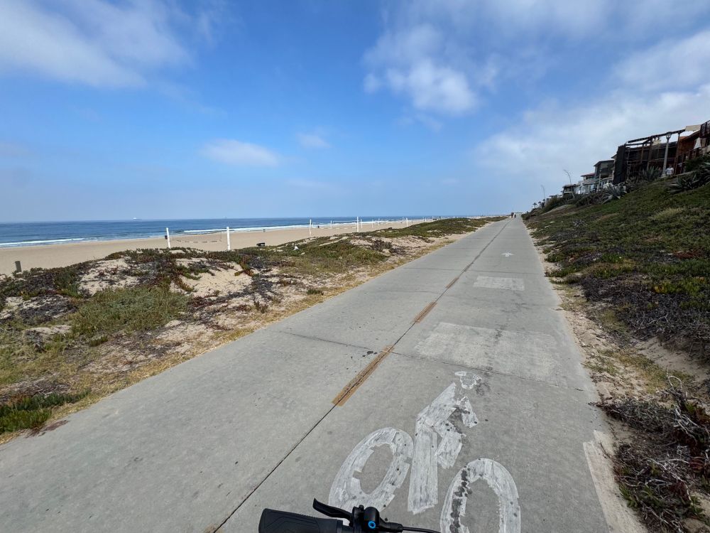A bike path along the beach in the Los Angeles suburb of Manhattan beach. The sky is blue, the sun is shining, and it looks like a perfect day.
There’s a bike handlebar just visible in the bottom of the frame