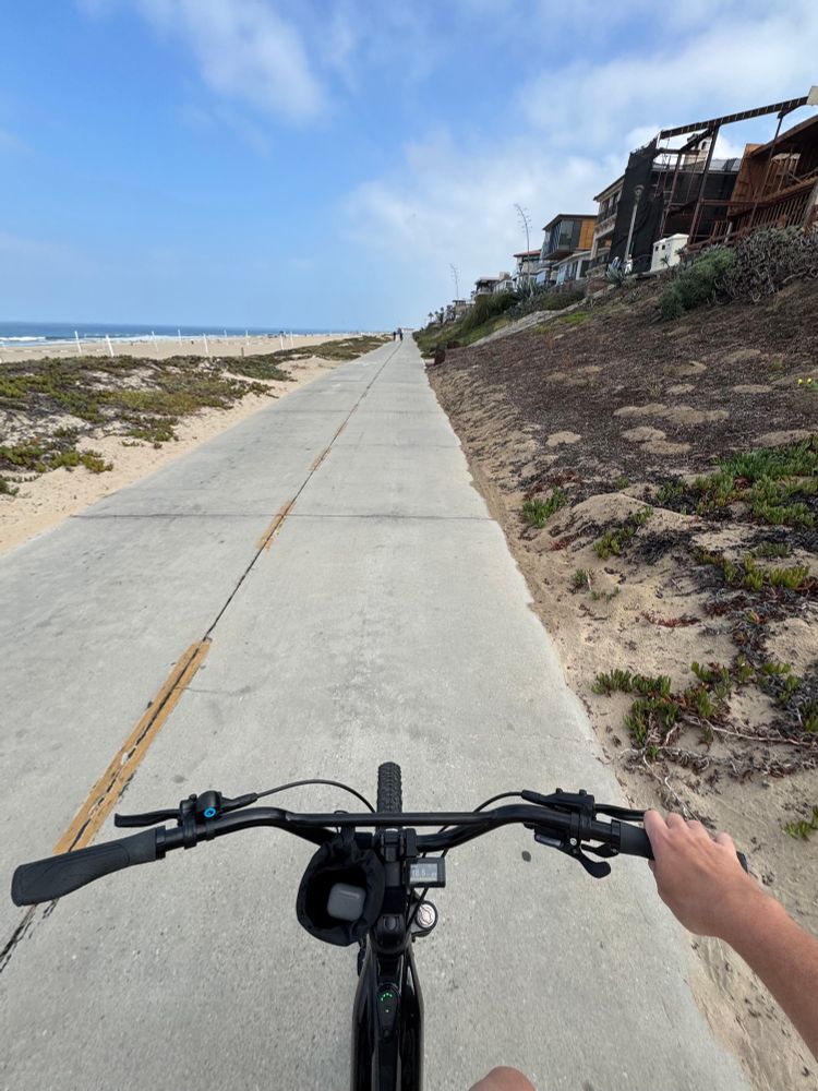 A bicycle cockpit aims down a beach bike path. It looks kinda like it belongs to a mountain bike, or perhaps an upright e-bike, or some tangled mess of the two. There’s a snack bag attached to the stem holding AirPods, and a hand holding the handlebars preventing the speed shakes from occurring.