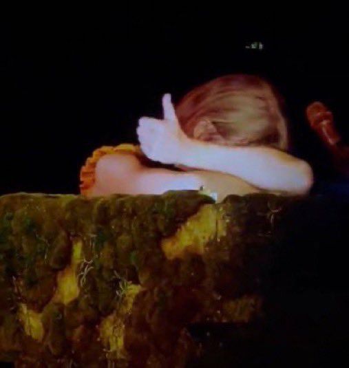 taylor during eras tour, seated at her moss-covered piano with her arms on the piano and her face tucked in there. she's giving a thumbsup. 