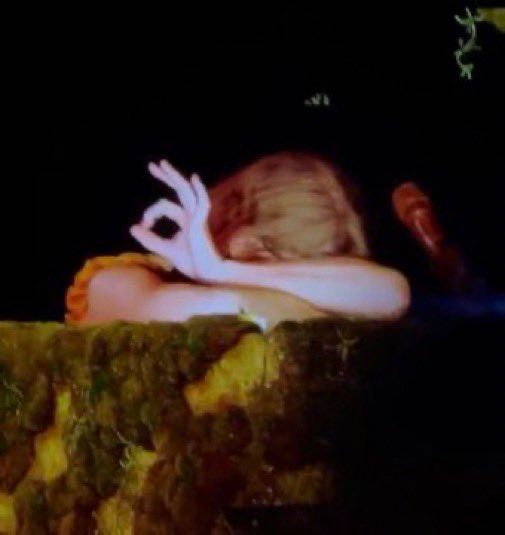 taylor during eras tour, seated at her moss-covered piano with her arms on the piano and her face tucked in there. she's giving an ok sign. 