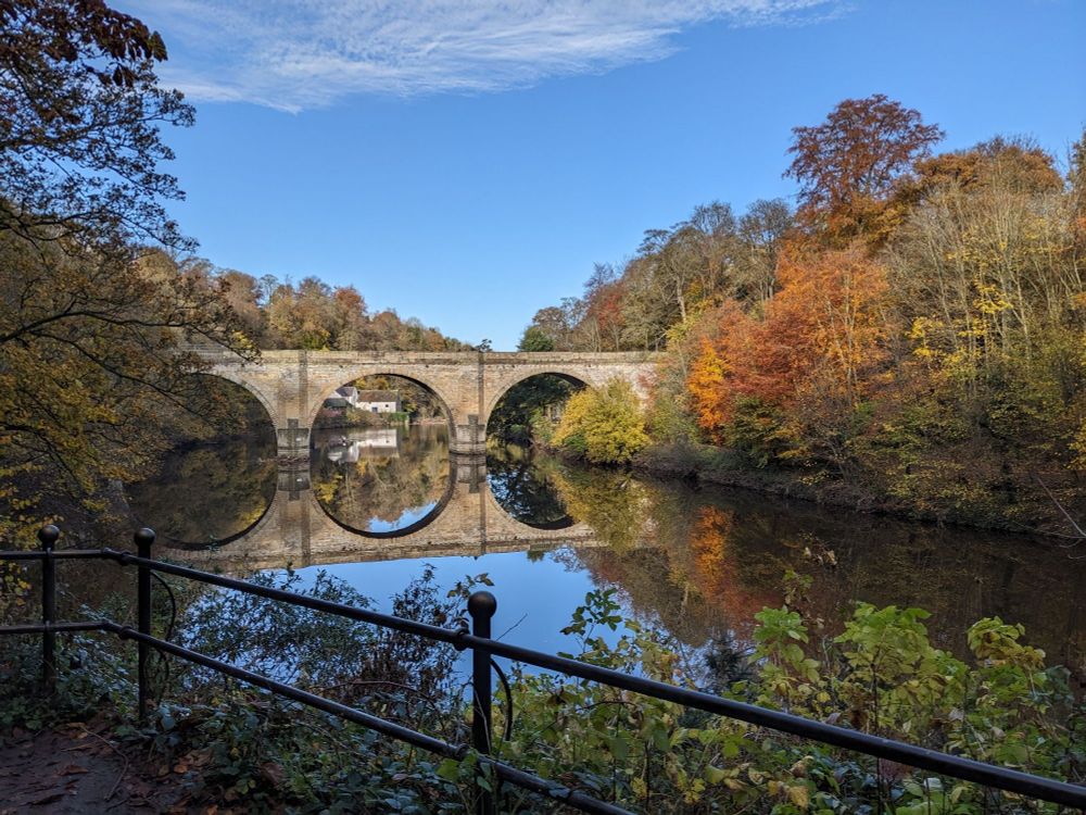 A photograph of Prebends Bridge in Durham (UK), with the River Wear's still waters in the foreground, autumn leaves on the trees lining the river, and clear blue skies above.