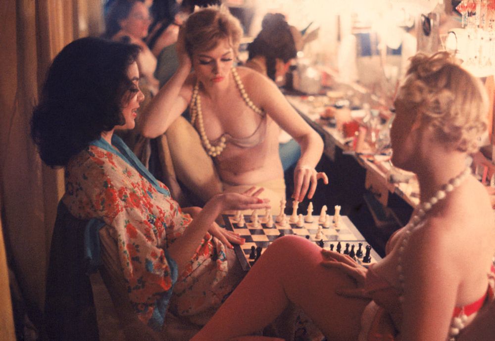 women in a dressing room, in various sates of dress, two of them are playing chess