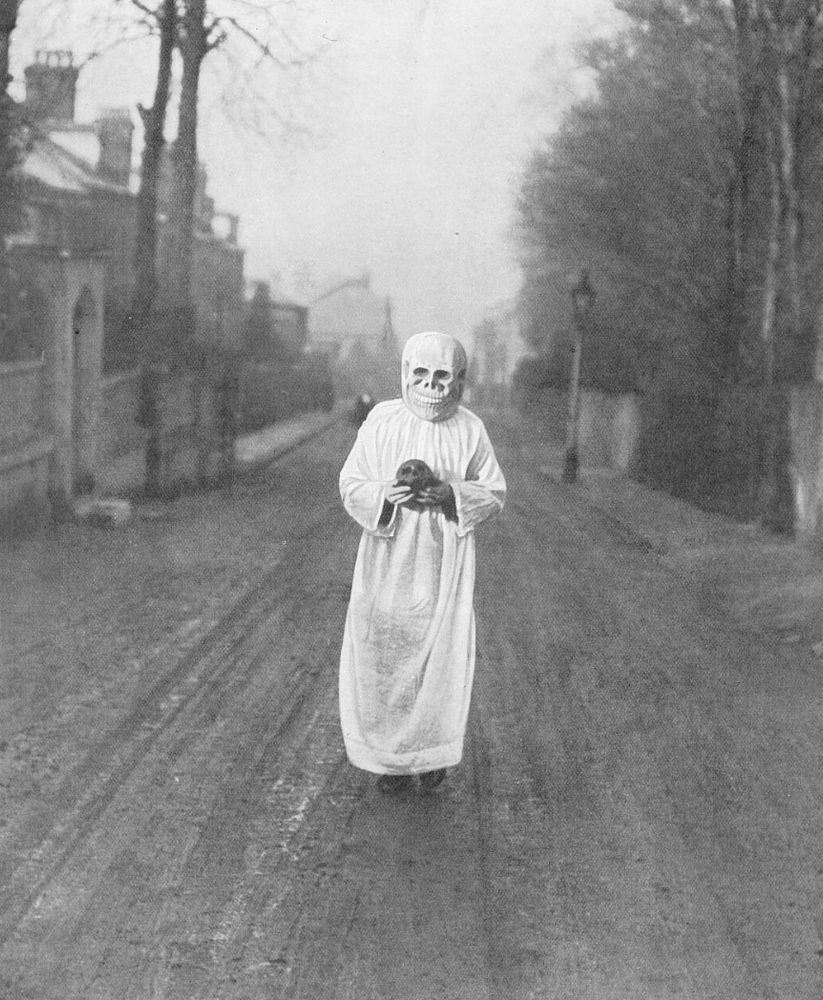 photo of a man walking up a street wearing a skeletal mask and a white robe, he is carrying a skull