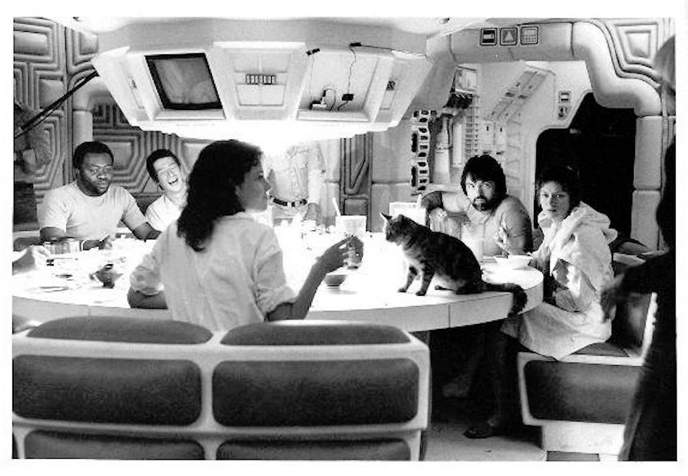 on set photo from Alien, 1979. The crew are sat around the table eating after being awoken, Jones is on the table begging, Jones is a cat.
