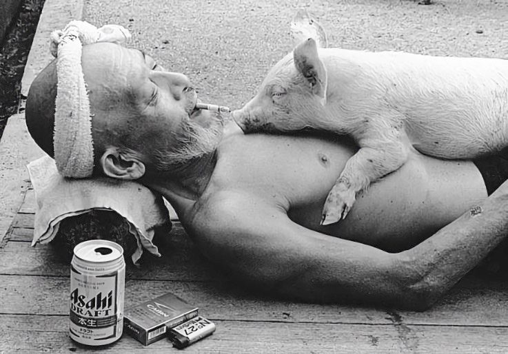 man lying down on a street, he is smoking, has a can of beer by his side and a sleeping pig on his chest.