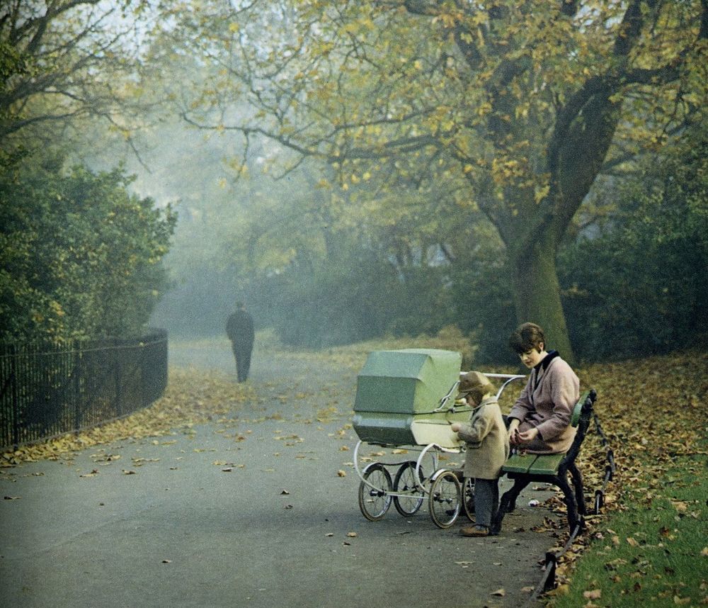 a misty park scene a woman with a pram sat on a bench. a figure is seen in the distance