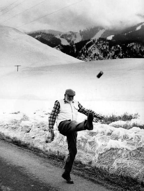 man in a snowy landscape, mountains in the distance. The man is kicking a can, his leg high in the air