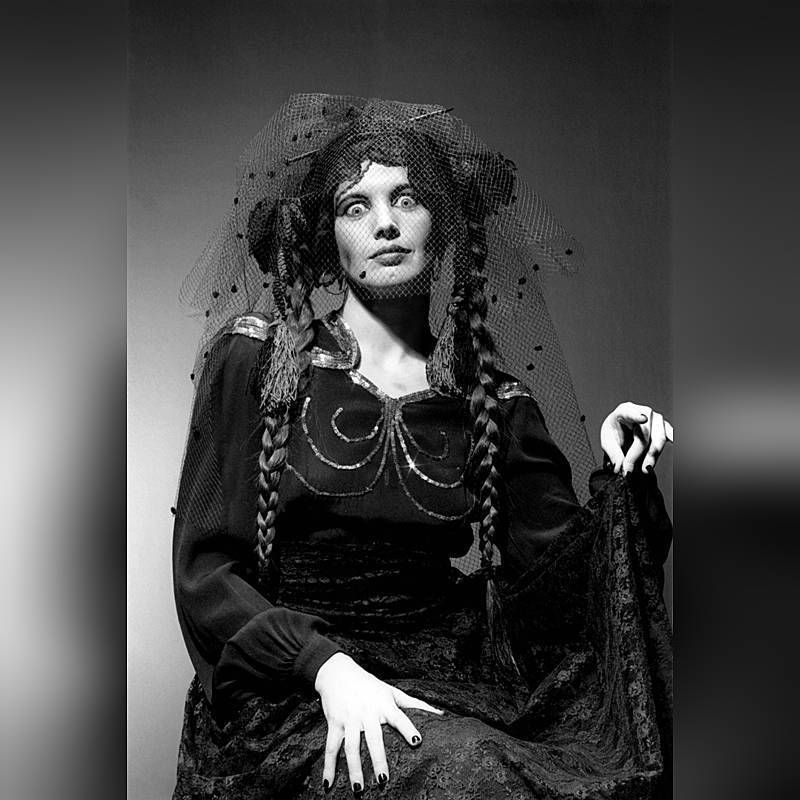 woman in a dark dress with a veil over her face, she is looking, wide eyed and is pointing with one hand.