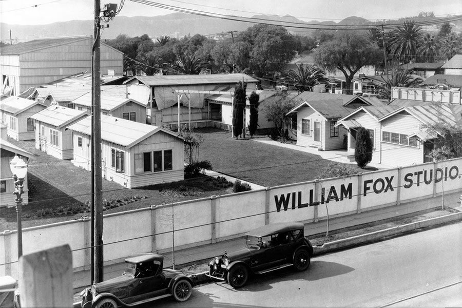 shot of some buildings enclosed by a fence., a sign reads william fox studios. a couple of parked cars on the road.