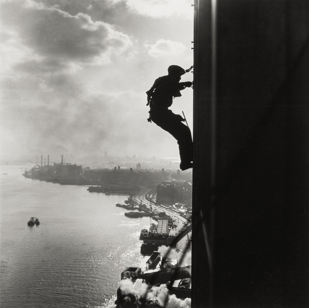 man washing windows on a tall building. below him is a city and a port.