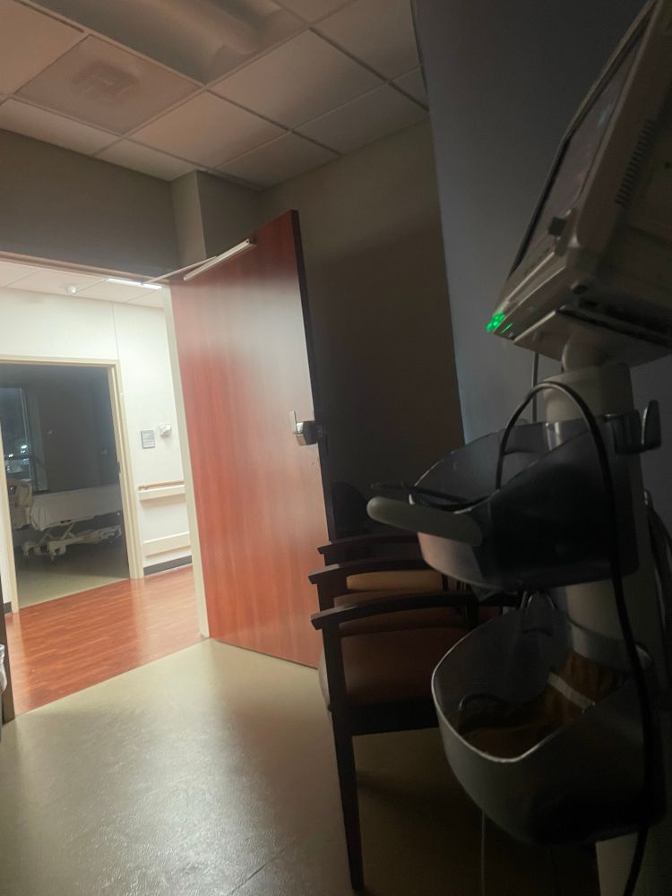 View from dark hospital room across the hall to another hospital room. Vital machine is to the right. Monitor is not visible. The door and the hall floor have weirdly similar wood designs and color