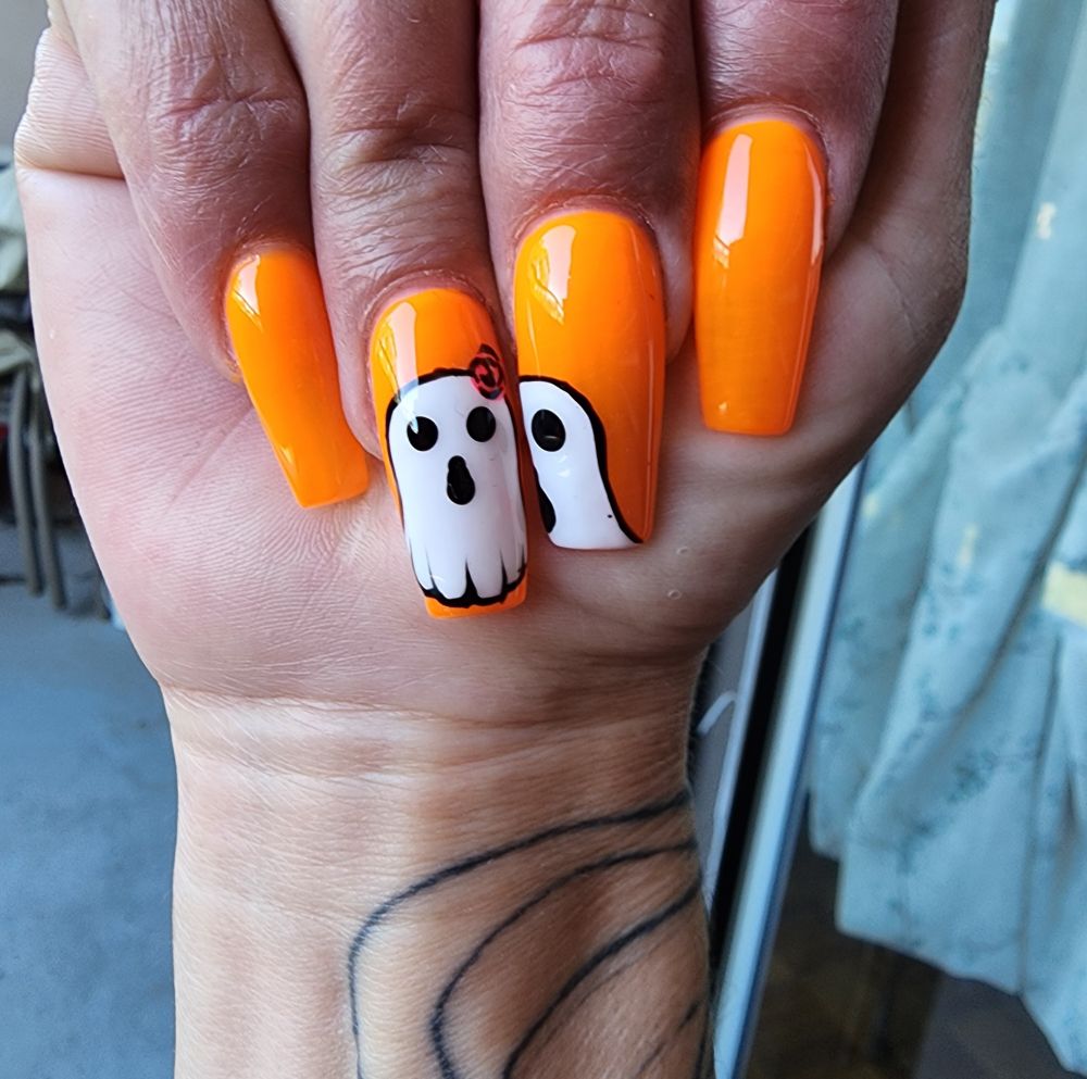 Image of a hand showing off some long coffin, orange nails with a couple of cute ghosts painted on the middle and ring fingernails. A tattoo is visible on the person's wrist. 