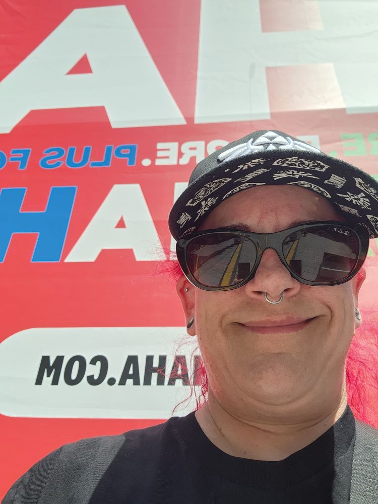 Image of a female presenting person, wearing a baseball hat, sunglasses, a black tshirt, and a smile. Said person is standing in front of a red sign with backwards lettering in white and blue letters