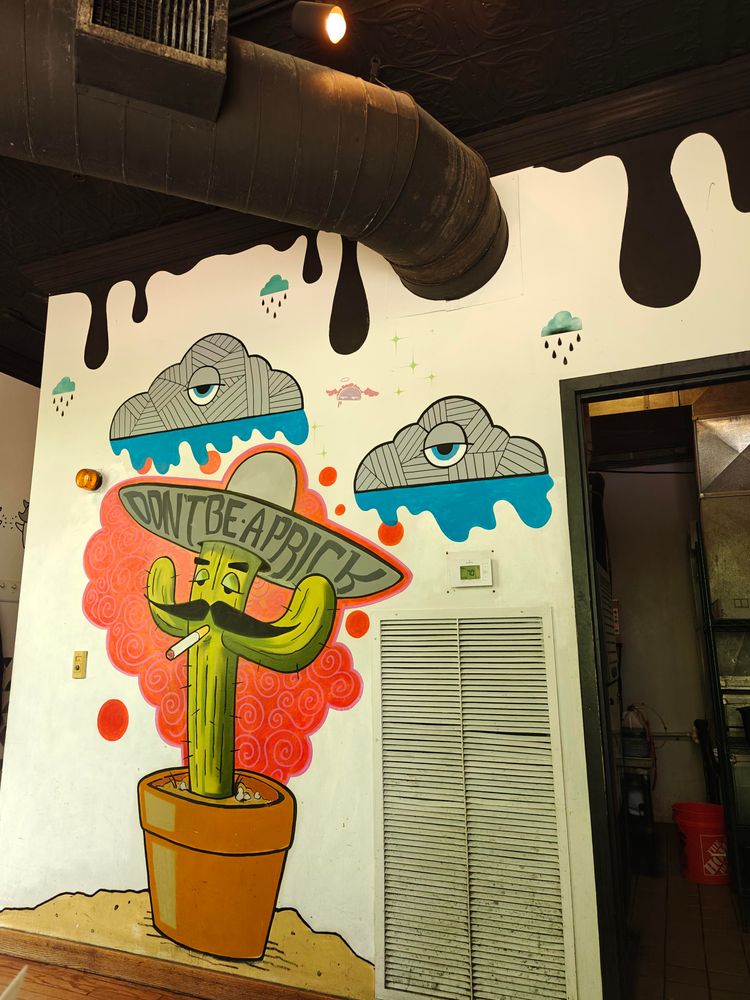 Mural of a cactus painting with a cigarette, mustache, and droopy eyes. Cactus is wearing a big sombrero with the words "don't be a prick"