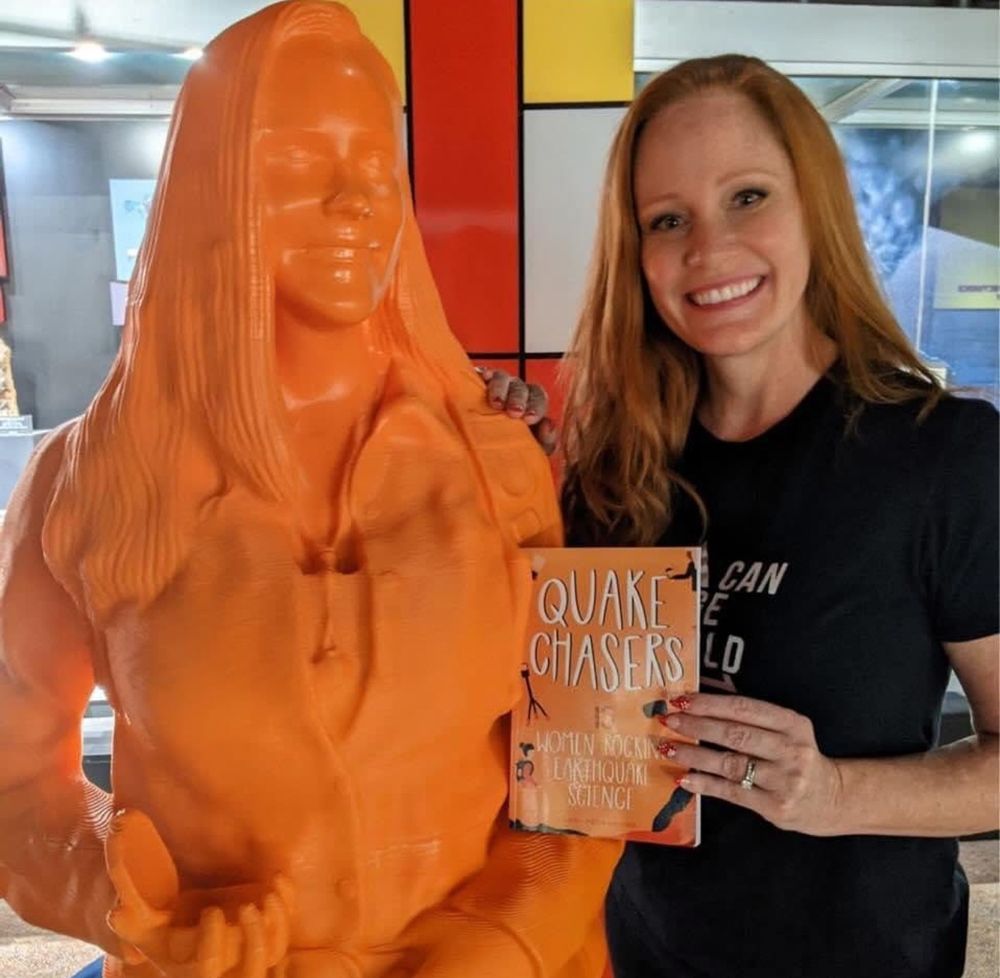 A smiling women stands next to an orange statue of herself holding a book titled “ Quake Chasers: 15 women shaking up earthquake science”