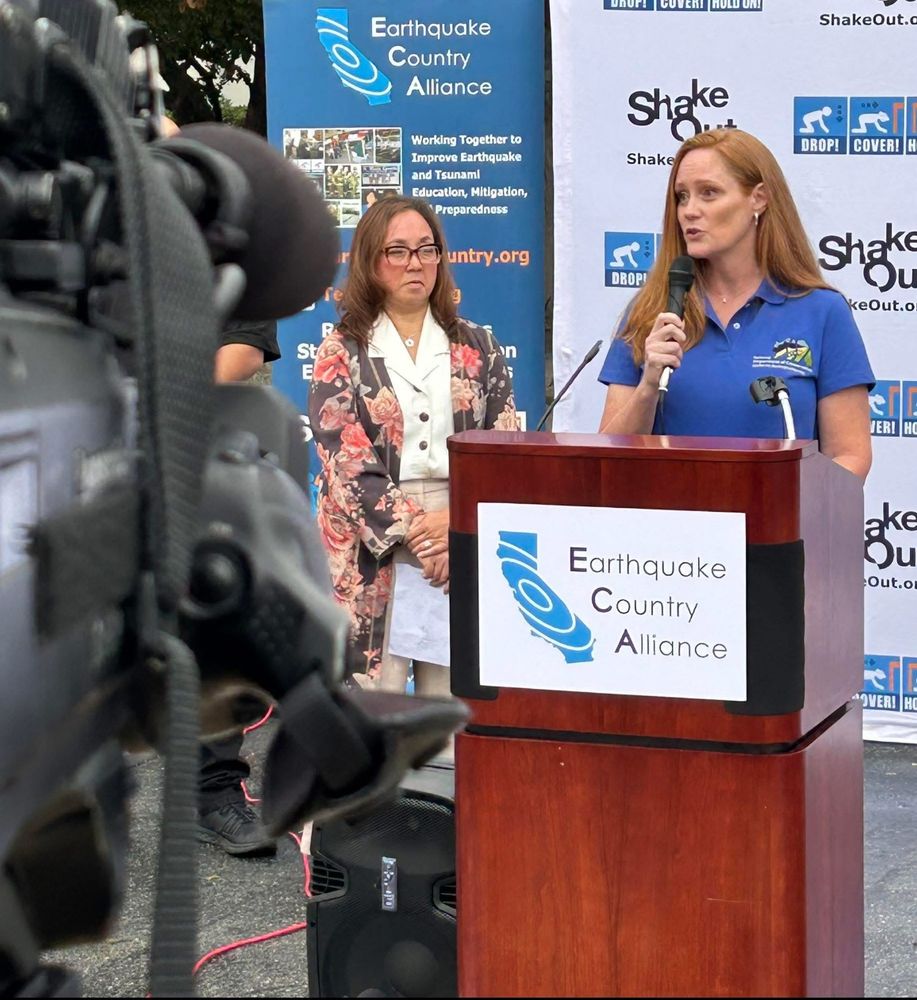 A women in a blue shirt stands behind a lecturn holding a microphone. A TV camera is in the foreground. 