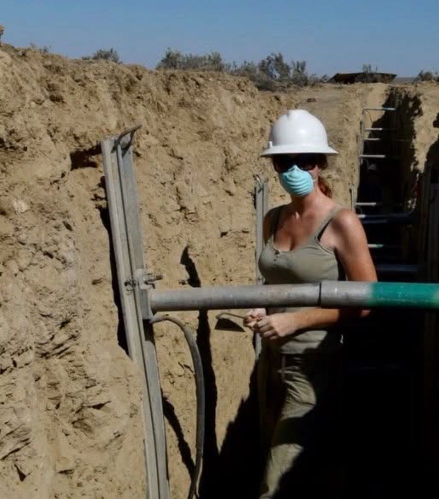 A women wearing a hardhat and mask stands in a paleoseismic trench holding a scrapping tool. 
