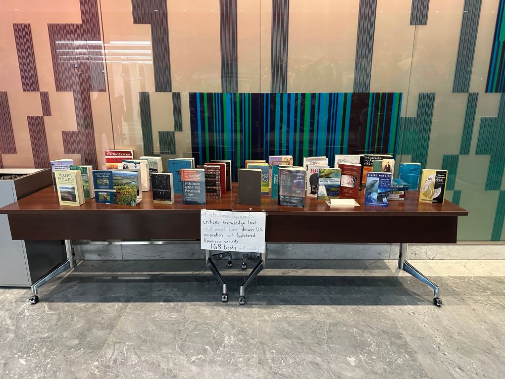 A table of books with a sign that says “each one of these books represents critical knowledge lost that would have driven US innovation and bolstered American security. 168 books and counting.