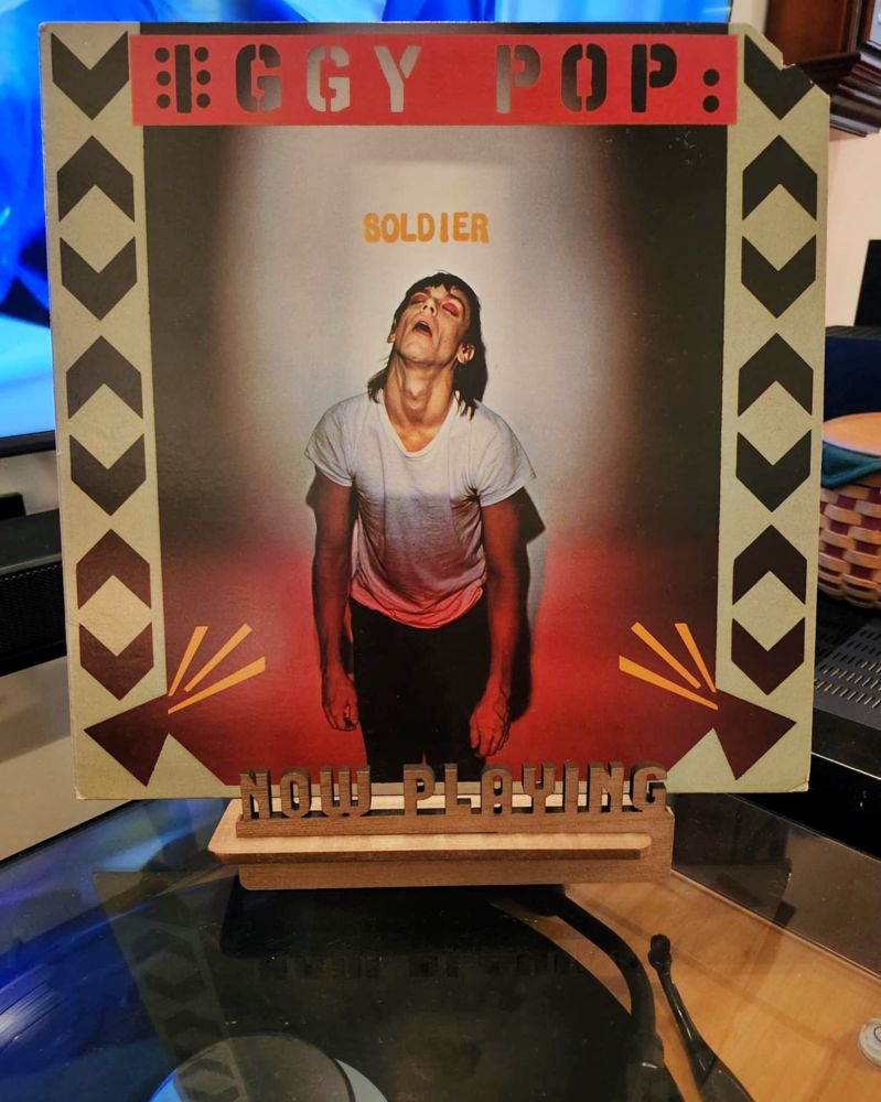 The front cover of the 1980 LP record “Soldier” by Iggy Pop sits on a wooden “now playing” record display stand placed on top of the turntable that’s playing the album. A muted television plays in the background.