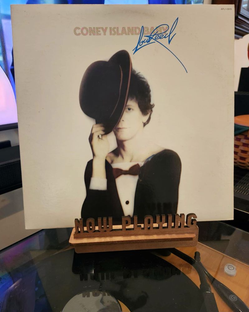 The front cover of the 1976 LP record “Coney Island Baby” by Lou Reed sits on a wooden “now playing” record display stand placed on top of the turntable that’s playing the album. A muted television plays in the background.