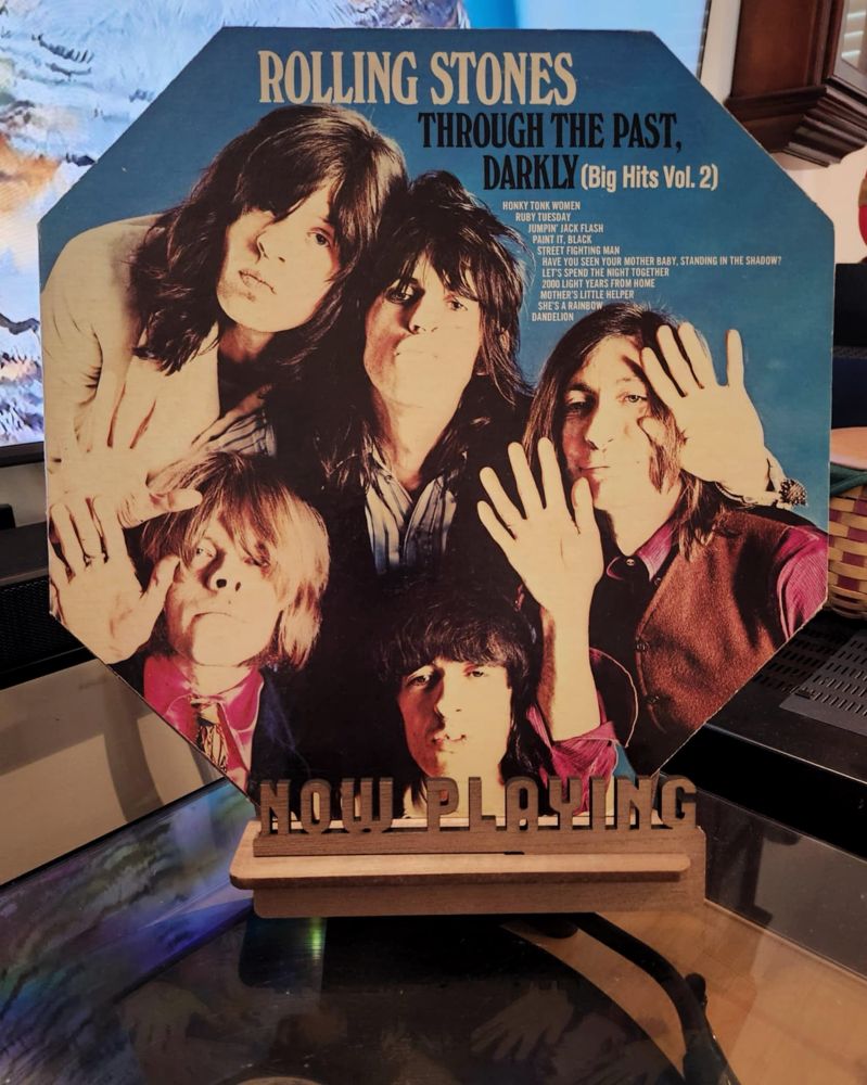 The front cover of the LP record “Through The Past, Darkly (Big Hits Vol. 2)” by The Rolling Stones sits on a wooden “now playing” record display stand placed on top of the turntable that’s playing the album.
A muted television plays in the background.