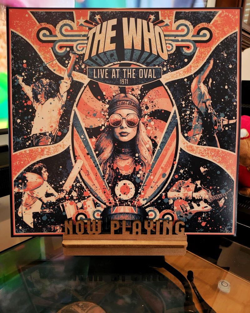 The front cover of the 2-LP live vinyl release by The Who, “Live At The Oval 1971”, sits on a wooden “now playing” record display stand placed on top of the turntable that’s playing the album. A muted television plays in the background.