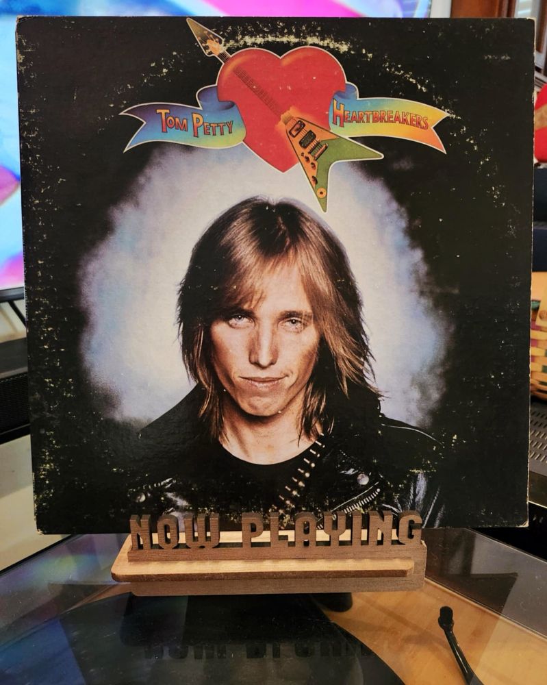 The front cover of the self-titled debut by Tom Petty and The Heartbreakers sits on a wooden “now playing” record display stand placed on top of the turntable that’s playing the album.
A muted television plays in the background.