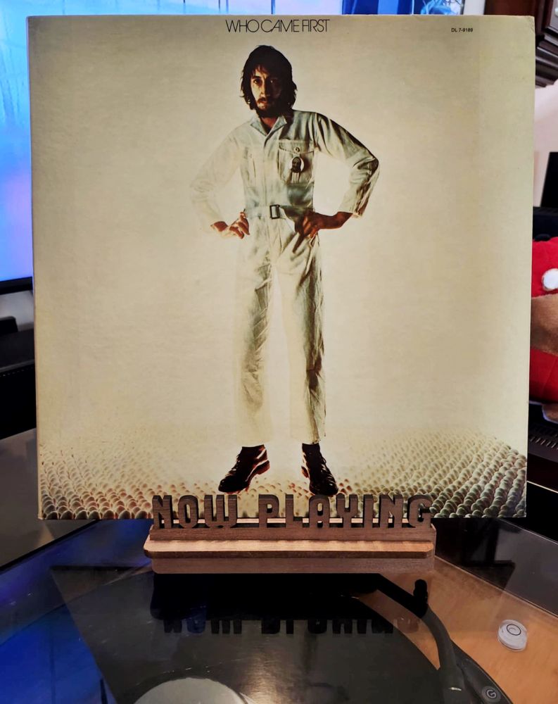 The front cover of the LP record “Who Came First” by Pete Townshend sits on a wooden “now playing” record display stand placed on top of the turntable that’s playing the album. A muted television plays in the background.