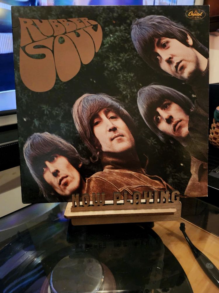 The front cover of the LP record “Rubber Soul” by The Beatles sits on a wooden “now playing” record display stand placed on top of the turntable that’s playing the album.
A muted television plays in the background.