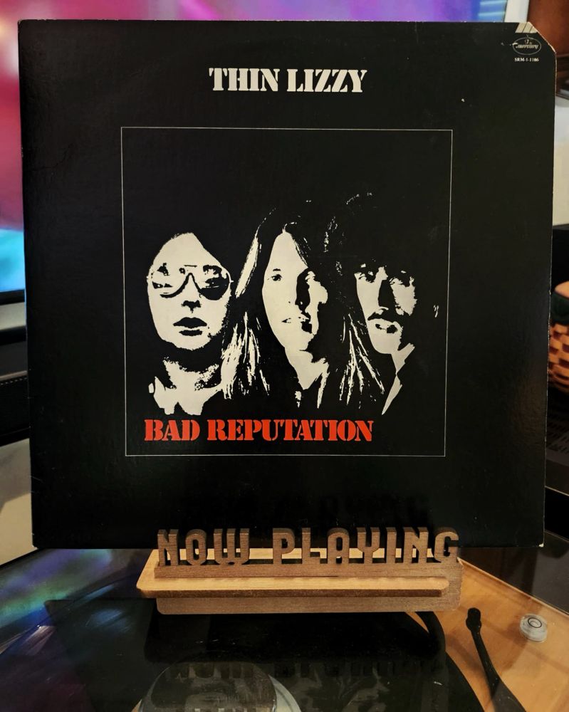 The front cover of the 1977 LP record ”Bad Reputation” by Thin Lizzy sits on a wooden “now playing” record display stand placed on top of the turntable that’s playing the album. A muted television plays in the background.